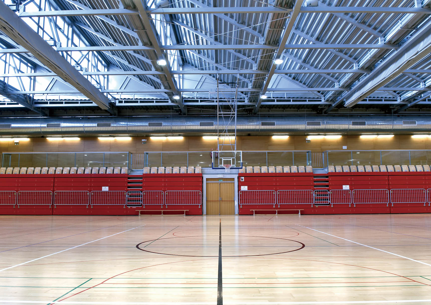 Empty sports hall with metal roof, red stand, central black line. Different colored lines lines on the floor define the court for different sports.